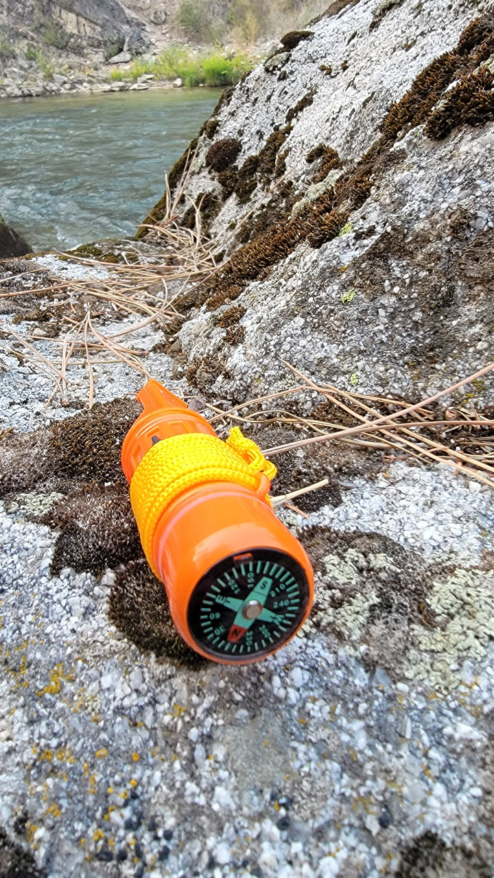 Orange 5-in-1 survival whistle with compass resting on rocks, essential gear for any bug-out bag and emergency preparedness.
